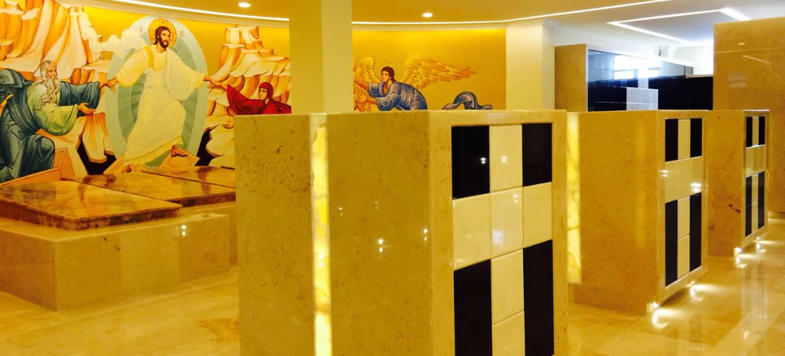 Criptas Espacio de mármol beige con diseño de nichos en blanco y negro. Al fondo, un mural religioso con figuras de Jesús y ángeles en tonos cálidos