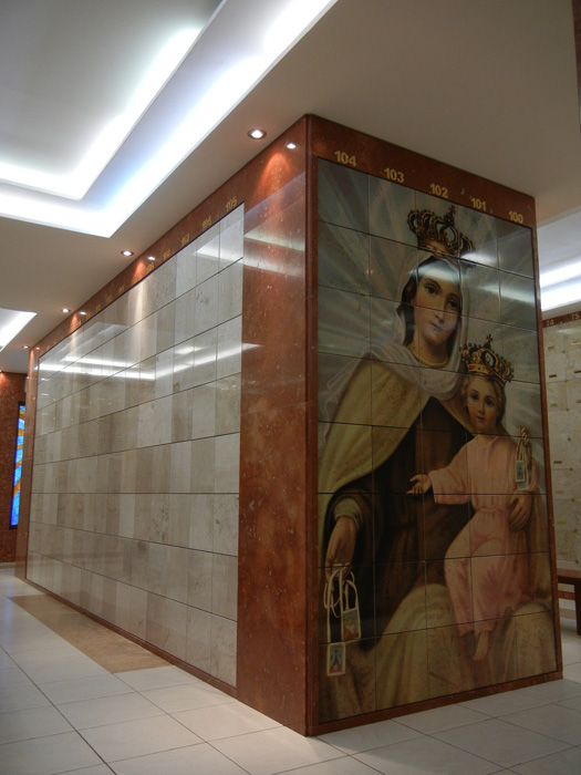 Criptas Imagen de pared de nichos de mármol con una ilustración de la Virgen del Carmen y el Niño en una esquina, numeración dorada en la parte superior y luz de techo indirecta.