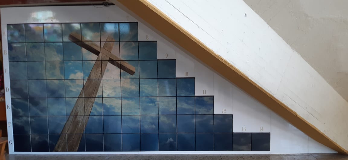 Criptas Mural en mosaico bajo una escalera, que muestra una cruz de madera contra un fondo de cielo, con numeración en los bordes