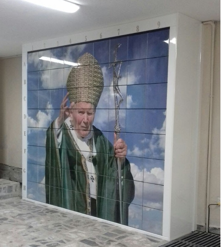 Criptas Pared interior decorada con una imagen de Juan Pablo II vestido con vestiduras verdes y mitra, sosteniendo un báculo pastoral, con un fondo de cielo azul con nubes. La imagen está dividida en secciones numeradas en los bordes superior e izquierdo
