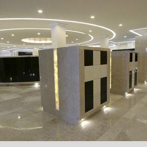 elegante espacio interior con columnas y estructuras revestidas en tonos claros y oscuros. El piso es de mármol pulido, reflejando la iluminación suave de luces empotradas y anillos luminosos en el techo, creando un ambiente moderno y solemn